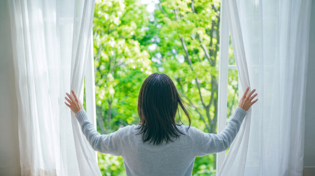 朝に窓を開けてカーテンを開け、新緑の景色を見つめる女性の後ろ姿。爽やかな暮らしやリフレッシュ、おうち時間をイメージしたライフスタイル写真