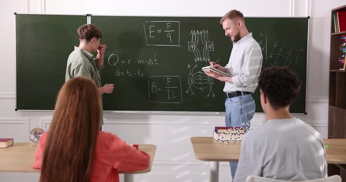 Student solving physics problems and teacher at chalkboard in classroom