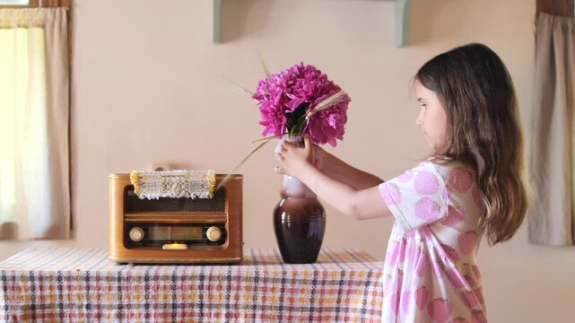 Young girl carefully arranging a vibrant bouquet of pink flowers in a vase beside an old wooden radio, evoking feelings of innocence, rustic charm, and a return to simpler times at home