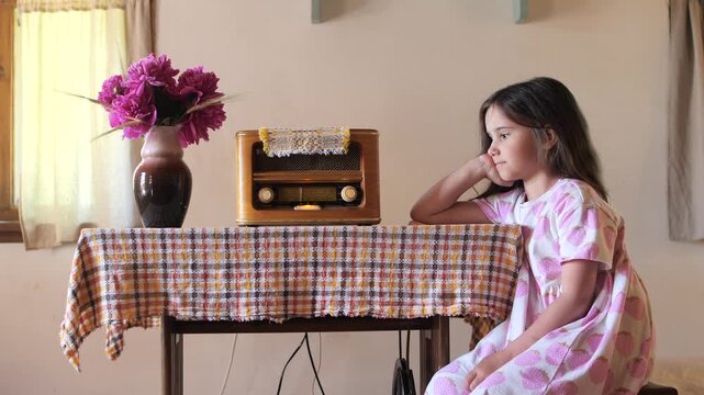 Young girl in a strawberry patterned dress resting her head on her hand. Sitting beside a classic wooden radio and a vase of pink peonies on a checkered tablecloth