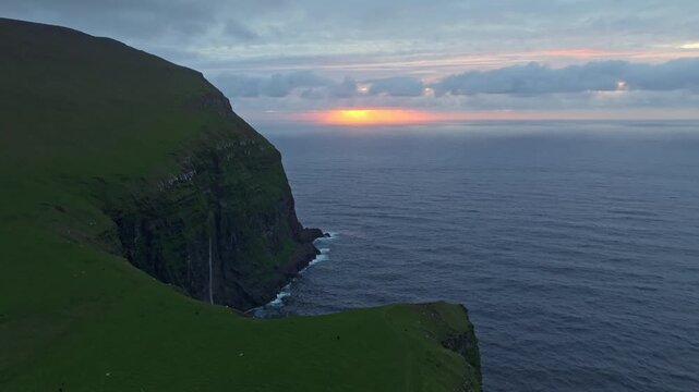 An aerial drone shot panning right shows a dramatic sunset over the sea cliff on the Faroe Islands. The vibrant colors of the sky meet the deep blue ocean, with a waterfall going down the  mountain.