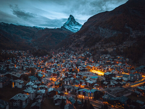 Aerial view of the Matterhorn towering over the illuminated town of Zermatt at dusk, featuring dense alpine architecture and mountain slopes in Zermatt, Valais, Switzerland.