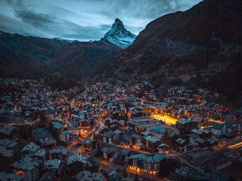 Aerial view of the Matterhorn towering over the illuminated town of Zermatt at dusk, with dense mountain chalets and glowing streetlights in Zermatt, Valais, Switzerland.