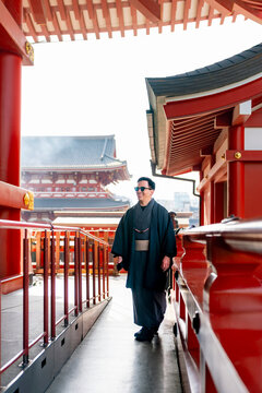 Young smiling Latin man walking relaxed through a temple corridor at Sensoji in Tokyo Japan, wearing a dark traditional kimono or yukata with elegant style, surrounded by vivid red wooden architecture
