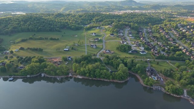 Aerial perspective of a suburban area in Johnson City, Tennessee, featuring residential houses, expansive green fields, and winding roads adjacent to a calm lake with distant hills in the background