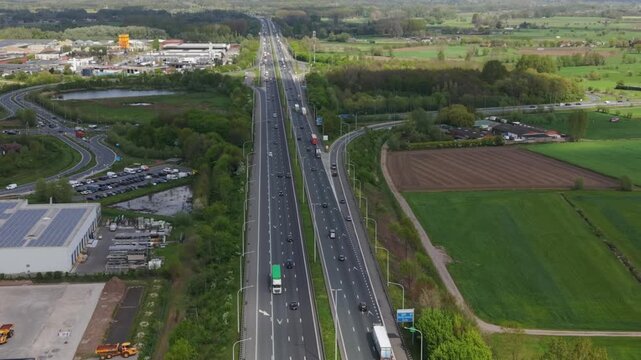 Aerial perspective captures a busy highway with vehicles traveling in multiple lanes, surrounded by green fields and industrial buildings, illustrating the dynamic flow of traffic in a rural landscape