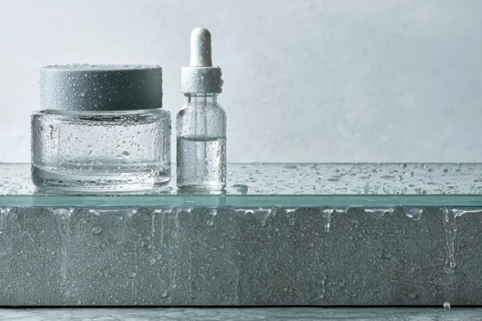 Clear glass skincare jar and dropper bottle on wet stone surface with water droplets