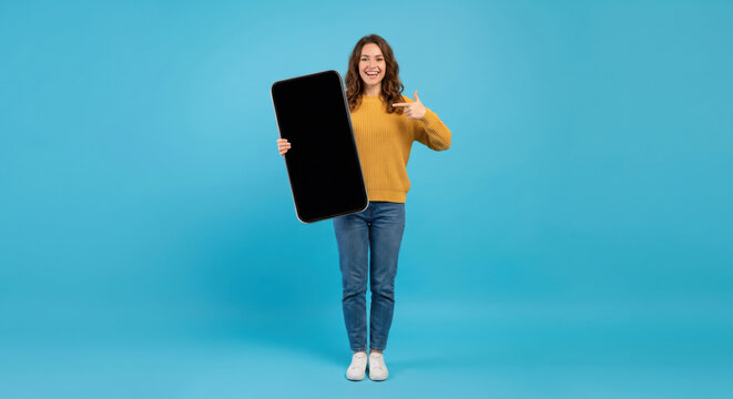 A person is standing on a blue background and holding a large black device. She is smiling and pointing at the device with her other hand. This scene takes place indoors
