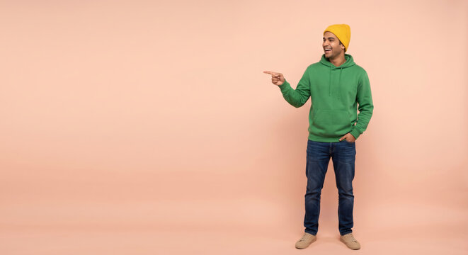 A man stands in front of a peach background. He wears a green hoodie and a yellow beanie. He smiles and points to the right with his left hand