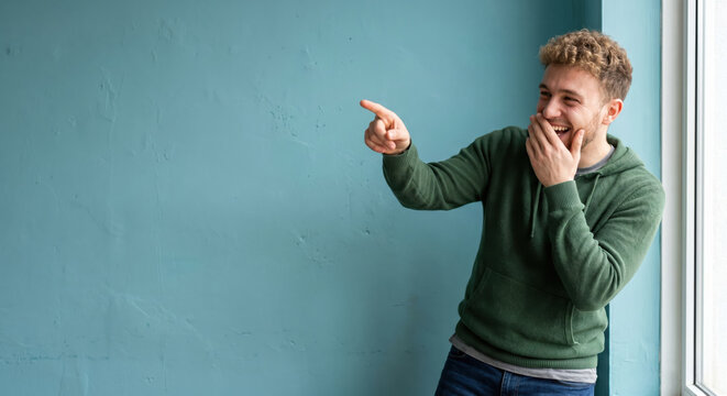 A man is smiling and laughing while pointing at something outside. He stands near a large window with a blue wall behind him. Sunlight comes through the window and brightens the room