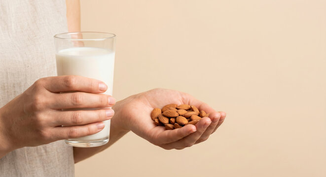 A person is holding a glass of milk in one hand while the other hand holds a handful of almonds. This scene takes place indoors against a plain background