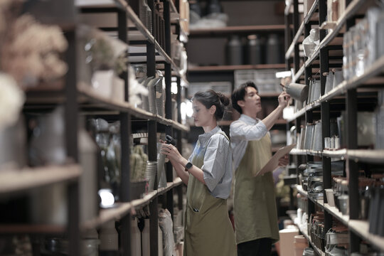 A young Asian craft shop owner in a product storeroom scanning items with a barcode reader during a stock count in a dimly lit stockroom. Handmade craft business.