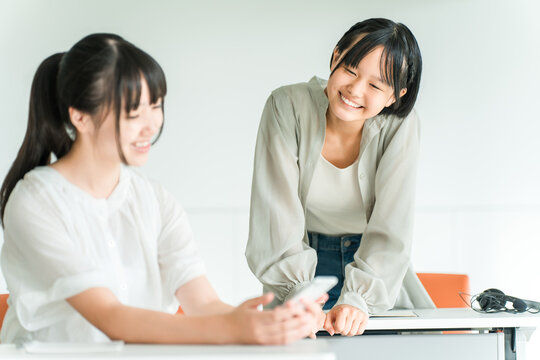 A female classmate is smiling and chatting in a classroom at the school/cram school, while holding her smartphone.