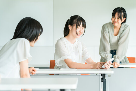 A female classmate is smiling and chatting in a classroom at the school/cram school, while holding her smartphone.