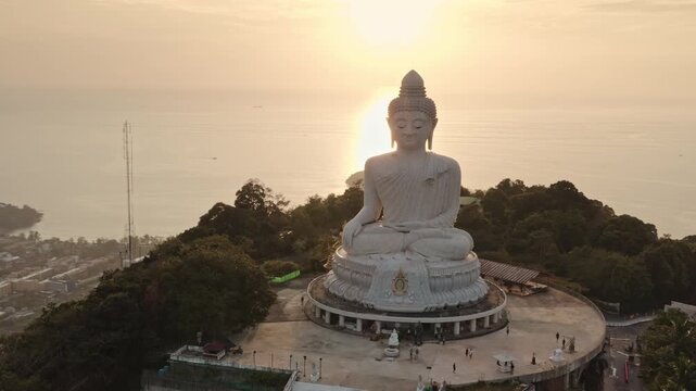 Big Buddha statue sitting on Nakkerd Hills, overlooking the sea with a vibrant warm light sunset creating a serene atmosphere and offering panoramic views of Phuket landscape. Aerial drone panorama