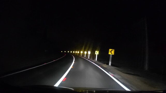 POV night drive along a winding rural road featuring reflective lane markings, curve warning signs and dark surroundings illuminated by vehicle headlights.