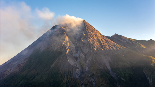 Aerial view of the volcanic crater of Mount Merapi with rising smoke and rugged rocky slopes illuminated by golden hour light in Balerante, Jawa Tengah, Indonesia.