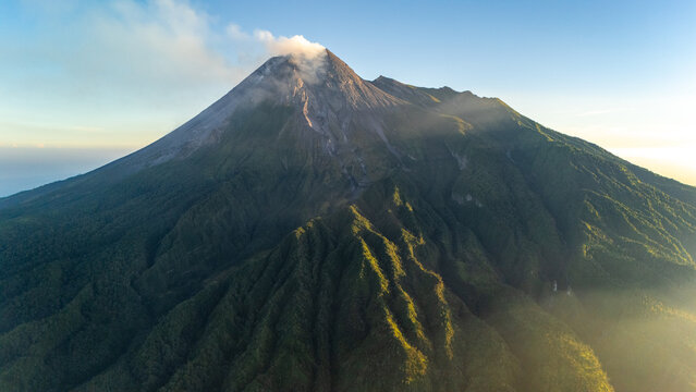 Aerial view of Mount Merapi with its volcanic crater emitting smoke, lush green forested slopes, and golden sunlight hitting the ridges Cangkringan, Daerah Istimewa Yogya, Indonesia.