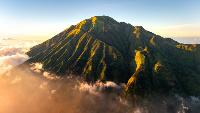 Aerial view of the lush green slopes of Mount Merbabu rising above a sea of clouds during golden hour in Selo, Jawa Tengah, Indonesia.
