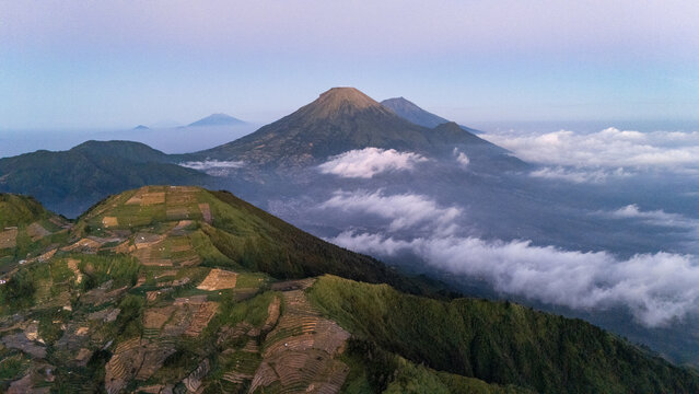Aerial view of the terraced agricultural fields on the slopes of the Dieng Plateau with Mount Prau in the background under a soft morning sky in Sembungan, Jawa Tengah, Indonesia.