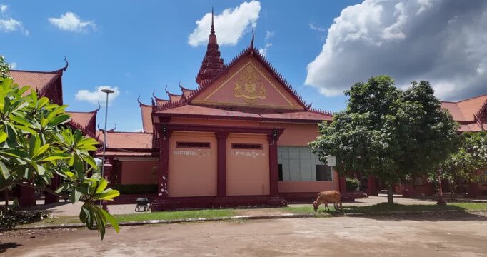 At the Cambodian border crossing, a traditional temple with an ornate reddish facade stands under a clear sky, while a cow grazes peacefully on the lawn in front of it.