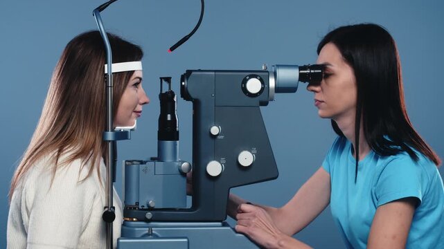Female optometrist conducts eye examination on patient using slit lamp in a clinical setting with blue background and medical equipment visible
