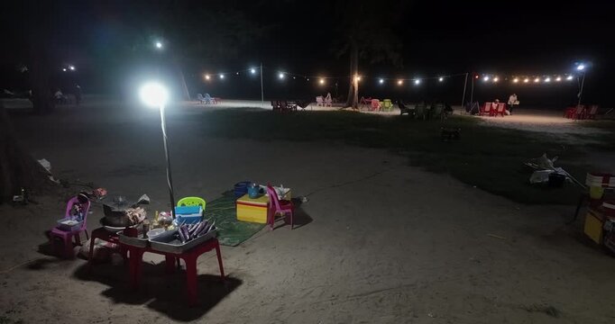 At night, a beach food stall with a bright lamp sits on the sand, while scattered groups of people relax at tables nearby, illuminated by a string of warm fairy lights hanging between the trees.