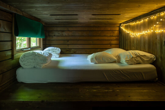 Intimate sleeping loft inside a wooden forest cabin with crisp white bedding and warm string lights glowing along the timber wall. A small green-blind window and soft amber light create a cozy