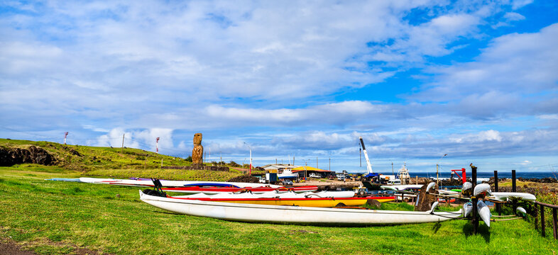 Outrigger canoes on green grass with the ancient Ahu Riata moai statue in the background at Hanga Piko harbor on Easter Island, Chile.