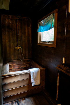 Dark wooden Japanese-style soaking tub with a vintage brass rain shower in a rustic forest cabin bathroom. A small window with a teal tie-dye blind overlooks the green forest and a white towel adds a
