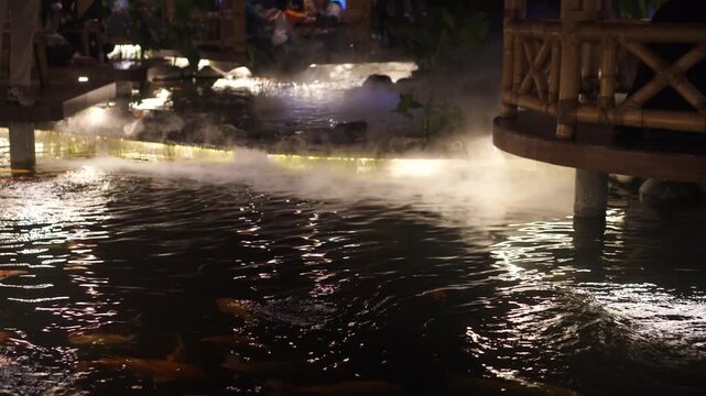 Ornamental koi fish swimming in a beautifully decorated pond beside a restaurant gazebo at night, illuminated by warm decorative lights and surrounded by rocks and gentle mist
