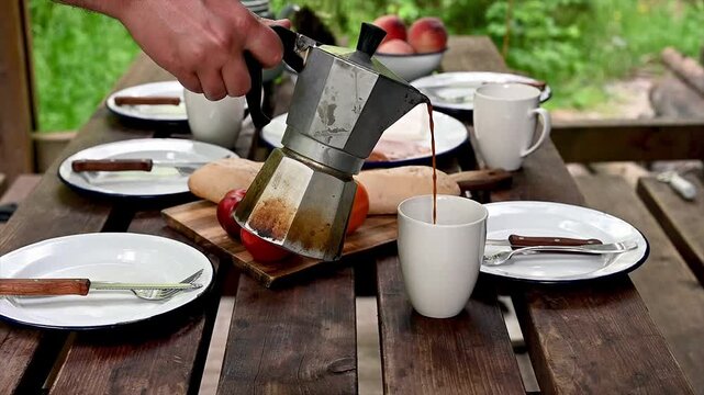 A hand pouring fresh coffee from a stovetop moka pot into an enamel mug at a rustic outdoor breakfast table with pancakes tomatoes and peaches. A cozy al fresco morning routine of camping coffee and