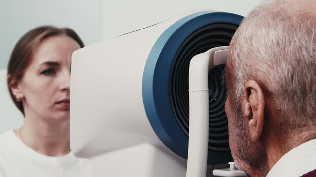 Elderly man undergoing computerized visual field examination with medical technician observing in a clinical setting, advanced equipment visible in the background