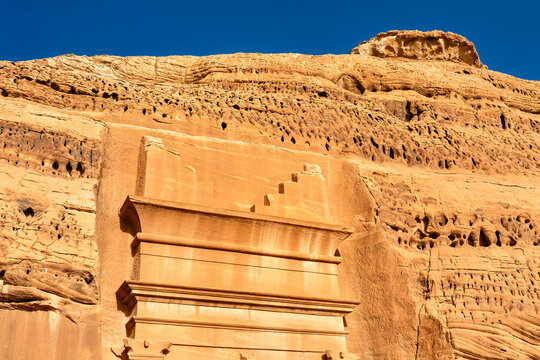 View of the intricate rock-cut tomb facade and sandstone cliffs at the Hegra archaeological site in Al Ula, Saudi Arabia.