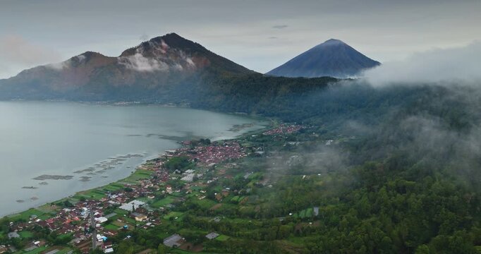 Aerial drone view showing the active Batur volcano caldera surrounding Lake Batur and Kedisan village with morning mist rolling over the lush green landscape and the distant Mount Agung peak, Bali