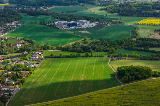 Aerial view of a logistics facility surrounded by green fields, agricultural land, and a small residential area in Kunice, Central Bohemian Region, Czechia.