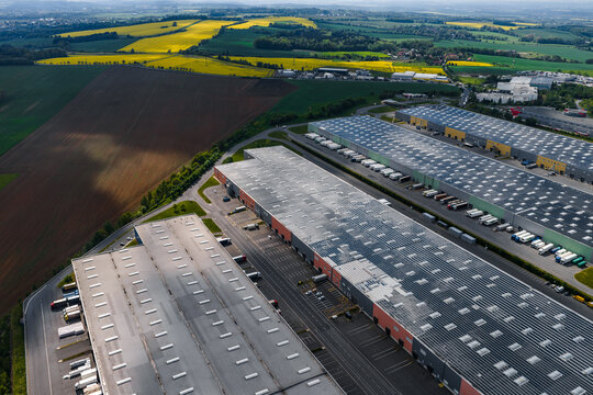 Aerial view of a large logistics center with warehouses and loading bays filled with trucks, surrounded by yellow rapeseed fields in Kunice, Central Bohemian Region, Czechia.