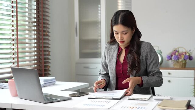 Professional young asian businesswoman analyzing business data and financial report while working at her modern office desk with a laptop and calculator