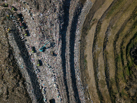 Aerial view of TPA Suwung landfill showing massive piles of waste, heavy machinery, and trucks operating on the site, with terraced slopes in Kecamatan Denpasar Selatan, Bali, Indonesia.