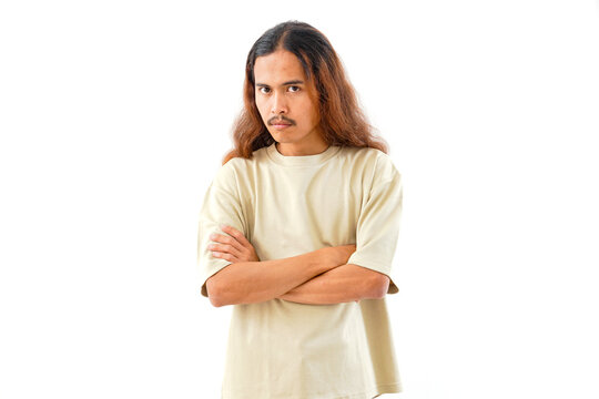 A studio portrait of a young man with long hair and a mustache, wearing a beige t-shirt, standing with his arms crossed and looking at the camera, isolated on white background.