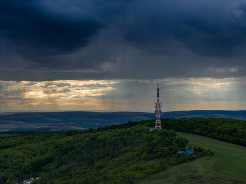 Aerial view of the telecommunications tower on a forested hill under a dramatic stormy sky with light rays breaking through the clouds in Strazovice, South Moravian Region, Czechia.