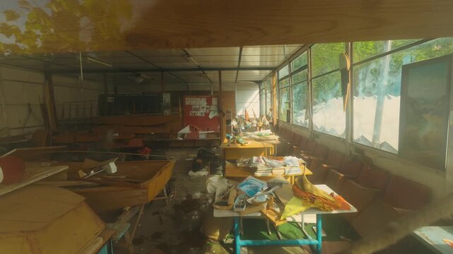 Abandoned school workshop interior featuring broken windows, scattered papers, wooden boat models, and debris. Sunlight streams through dust filling room creating eerie atmosphere
