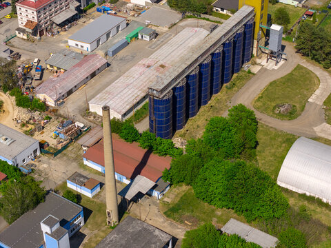 Aerial view of an industrial facility featuring a row of tall blue silos, a brick chimney, and surrounding warehouse buildings in Slavkov u Brna, South Moravian Region, Czechia.