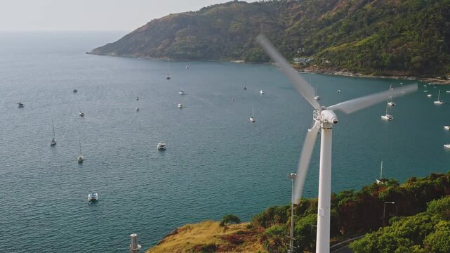 Wind turbine generating clean energy from the wind, overlooking a bay filled with sailboats and yachts anchored in the peaceful turquoise sea near the lush green coastline of Phuket island, Thailand