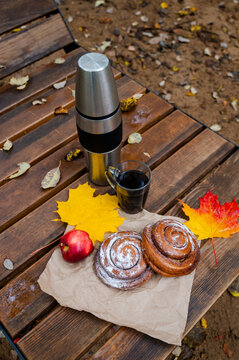 Autumn picnic setup with thermos, coffee mugs, cinnamon rolls, apple, and yellow maple leaves on a wooden bench outdoors