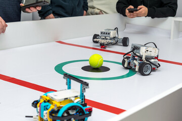 Children controlling robots via smartphones during a robot soccer game. High-angle shot of a white gaming table where several plastic DIY robots compete for a tennis ball. Kids in the background hold 