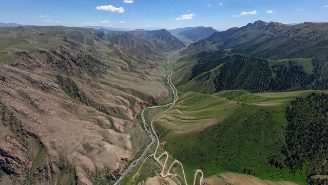 Aerial view of the winding mountain road leading through the lush green valley near Song-Kul, with a river flowing alongside rocky slopes under a clear blue sky in Naryn Region, Kyrgyzstan.