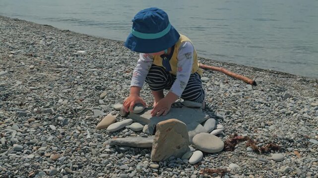Toddler in blue hat crouching on pebble beach arranging flat stones into structure. Calm water background. Natural daylight. Soft colors. Playful mood