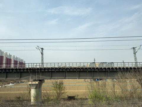 Railway bridge with power lines crosses fields near apartment blocks under blue sky in Baoding City, China