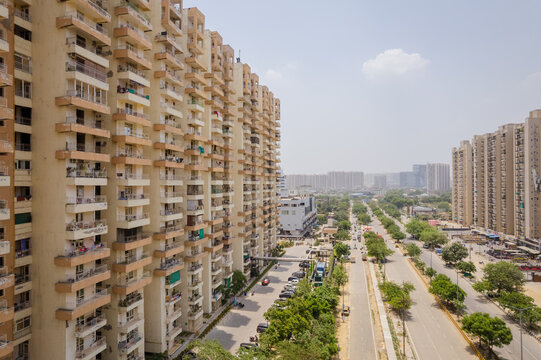 Aerial view of high-rise residential apartment buildings lining a wide road with lush green trees and urban infrastructure in Greater Noida, Uttar Pradesh, India.